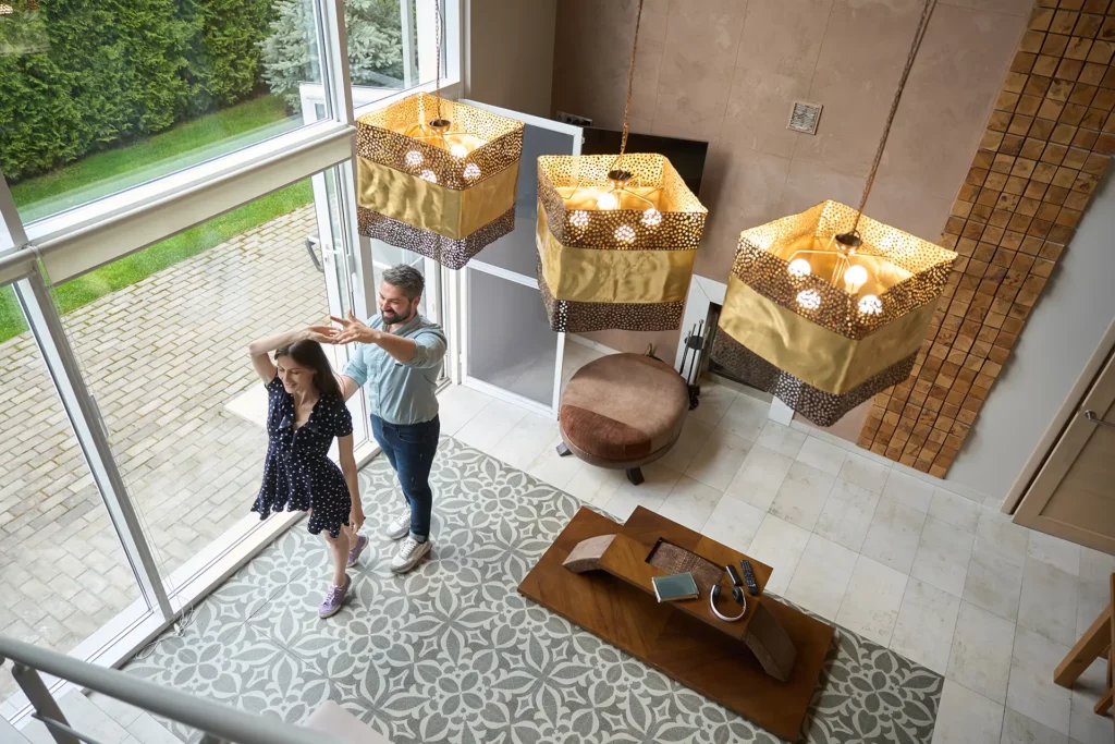 Couple dances in the modern living room with large windows, geometric rug, wooden coffee table, and stylish lights.